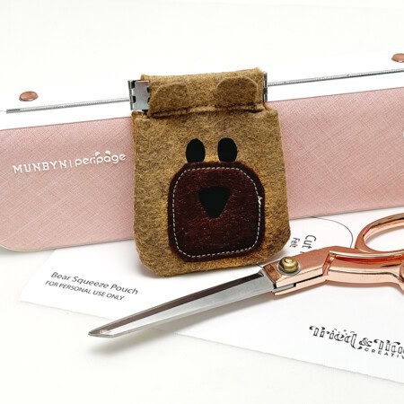A small felt bear coin purse, placed on a table with a pair of scissors and a pink Munbyn Peripage printer in the background. The pouch has a label reading "Bear Squeeze Pouch - For Personal Use Only.