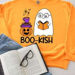 Bright orange sweatshirt with a cute ghost reading a book, a pumpkin, and a stack of books with a cup on top. Text reads "BOO-KISH." Open book and glasses next to it.