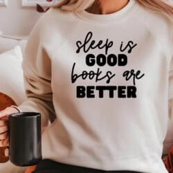 Person wearing a sweatshirt with the text "sleep is good, books are better," holding a black mug.