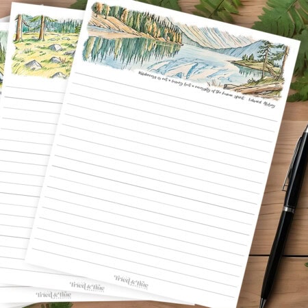 Two blank lined notepad sheets with nature-themed headers featuring landscapes and a quote from Edward Abbey. Perfect as camping journal pages, a black pen is placed beside the sheets, ready to capture your outdoor adventures.