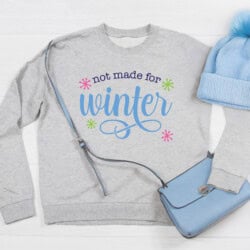 Gray sweatshirt with "not made for winter" text, blue handbag, and a blue pom-pom beanie on a white surface.