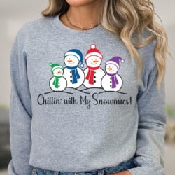 Woman wearing a gray sweatshirt featuring a snowman family graphic and text that reads "Chillin' with My Snowmies!.