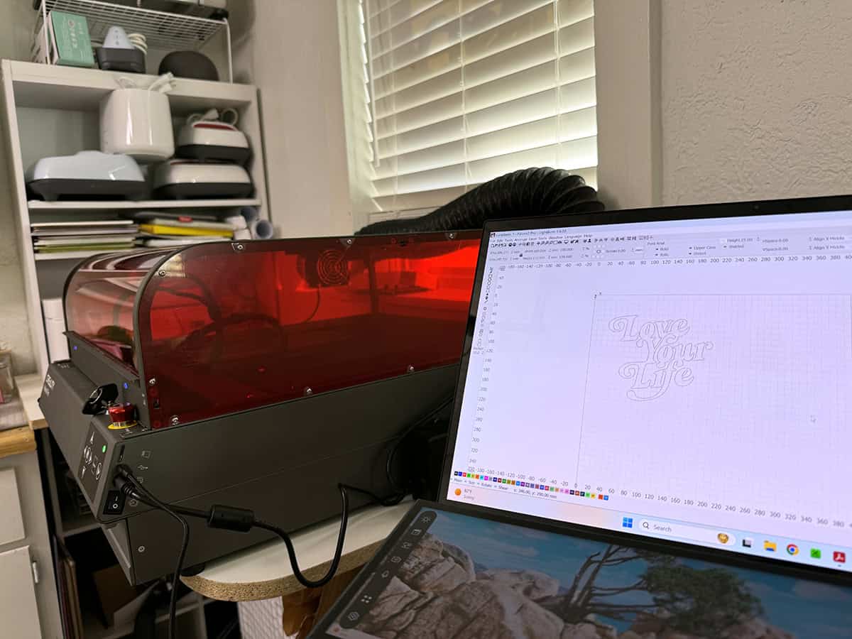 A 3D laser printer on a desk next to a computer screen displaying a design with the words "Love Your Life." The setup is in a room with shelves and a window with blinds.
