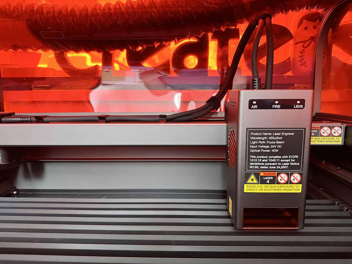 Close-up of a laser engraver's control unit inside an industrial machine, with caution labels and power cords. The background has an orange-red glow, likely from protective shielding.