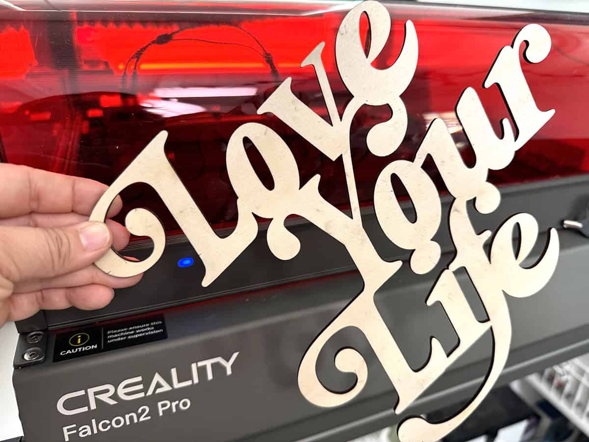 A hand holding a wooden cutout with the words "Love Your Life" in front of a Creality Falcon2 Pro laser engraving machine.