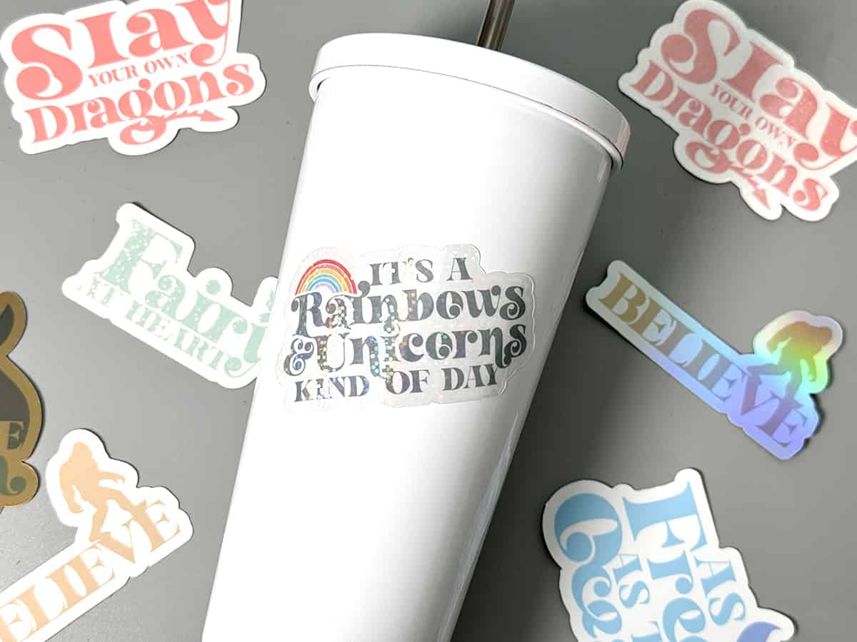 A white tumbler with the phrase "it's a rainbows & unicorns kind of day" surrounded by various colorful stickers with motivational and whimsical phrases.