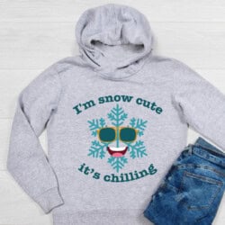 Gray hoodie with a smiling snowflake wearing sunglasses and the text "I'm snow cute, it's chilling." Beside it, folded blue jeans.