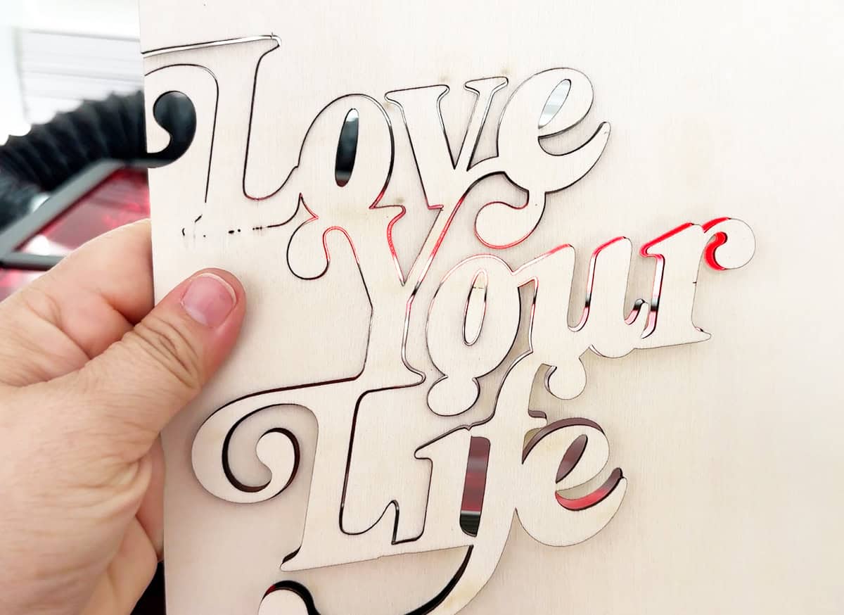 A hand holds a wooden piece with the words "Love Your Life" intricately cut out.