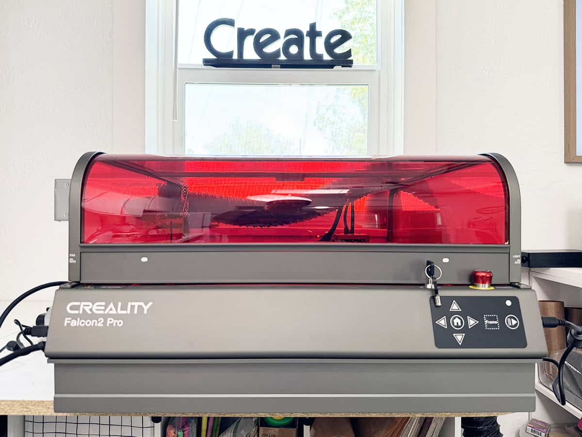 A Creality Falcon2 Pro laser cutter is on a table beneath a window with a "Create" sign. The machine has a transparent red cover and a control panel on the front.