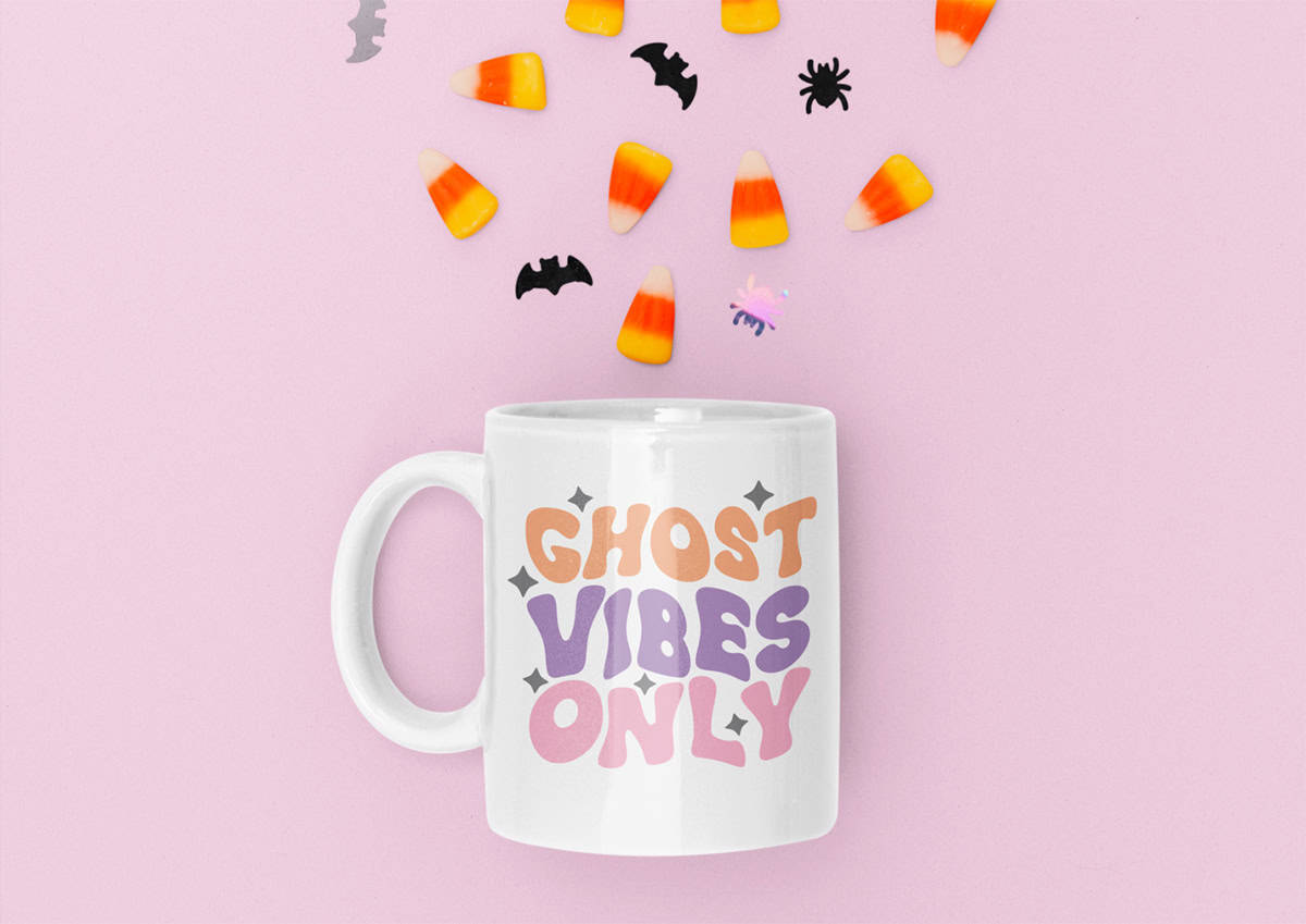 A white mug with "Ghost Vibes Only" in pastel colors is placed on a pink background with scattered candy corn, bat, and spider decorations.