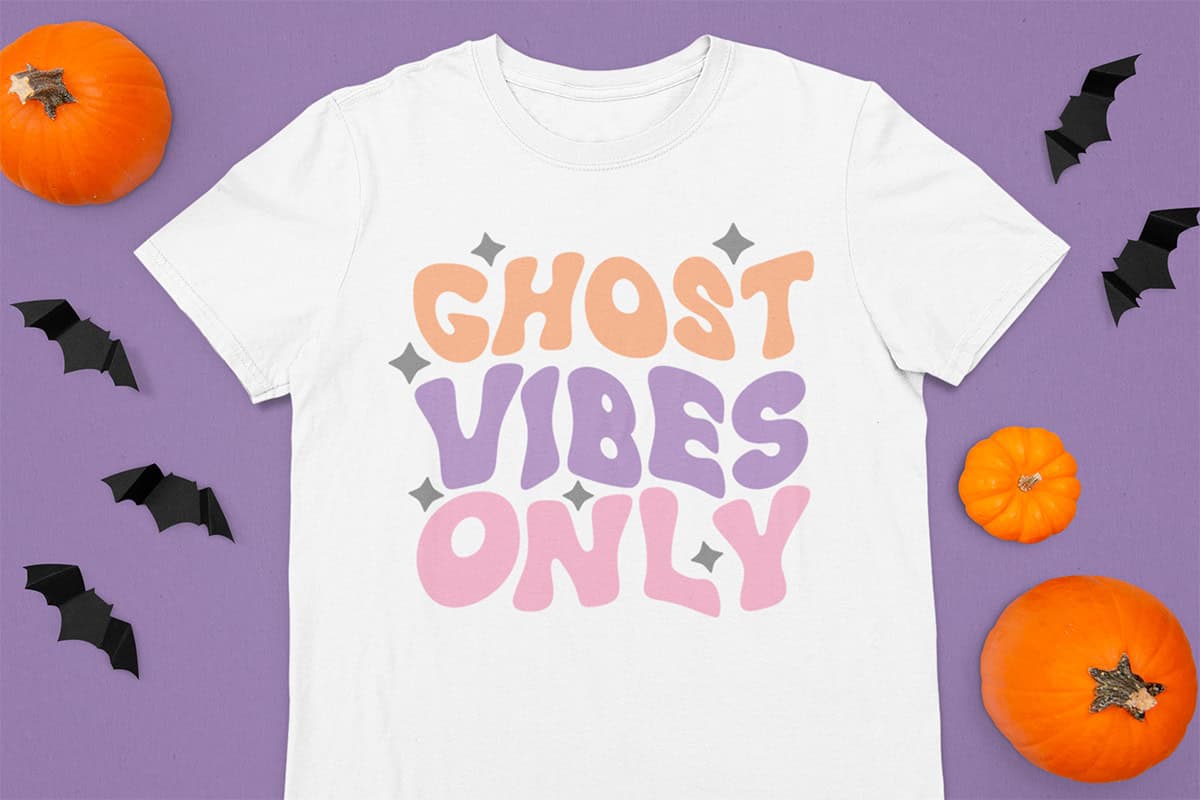White T-shirt with "Ghost Vibes Only" in orange, lavender, and pink letters. Surrounded by small black bats and orange pumpkins on a purple background.