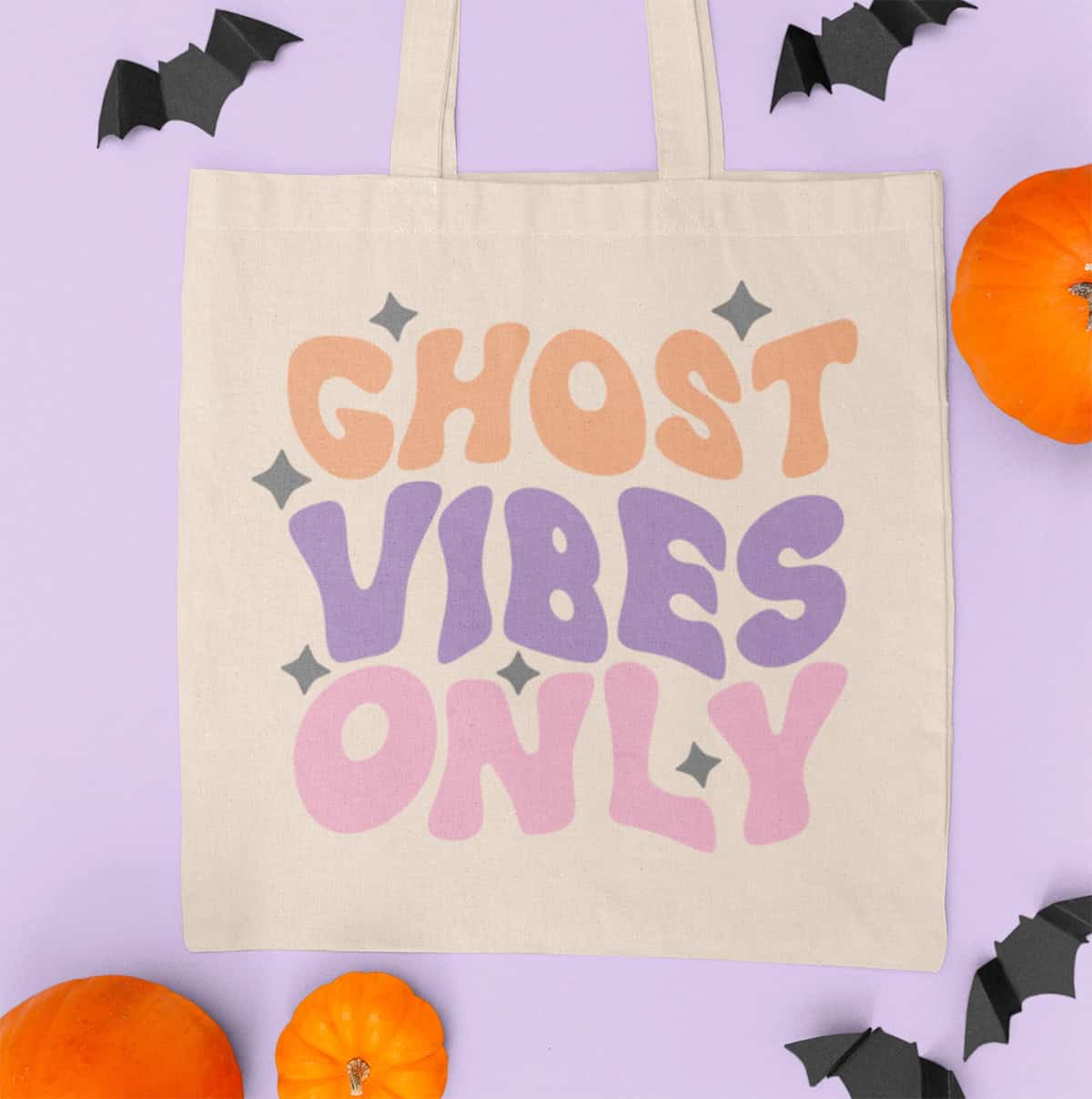 A beige tote bag emblazoned with "Ghost Vibes Only" in orange, purple, and pink delivers groovy Halloween flair. Adorned with black bats and pumpkins against a light purple backdrop, this accessory perfectly meshes festive spirit with style.
