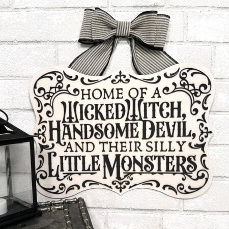 A charming Halloween family sign with decorative text reads, "Home of a wicked witch, handsome devil, and their silly little monsters," hangs on a white brick wall with a bow at the top. Free SVG available for download.
