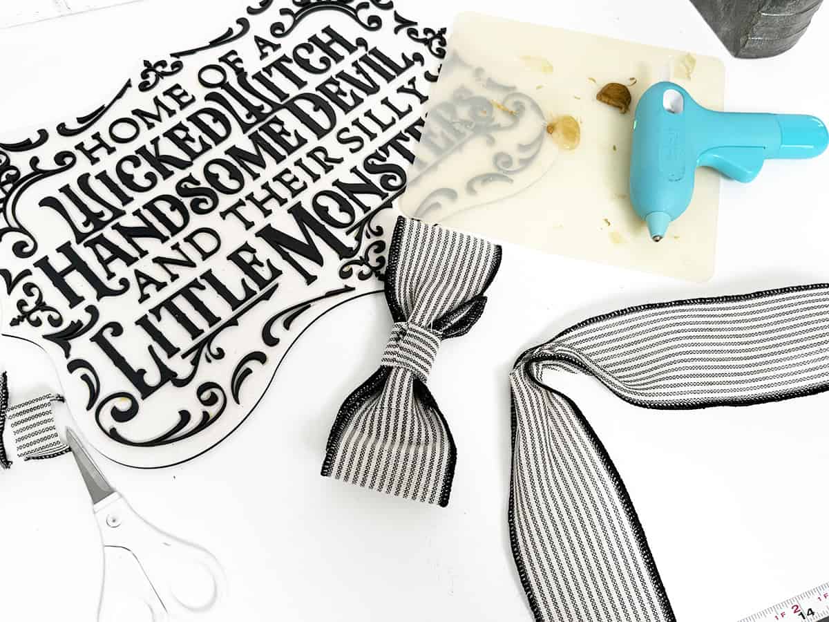 A craft project featuring a decorative sign with gothic text, a striped ribbon bow, scissors, a hot glue gun, and a partially used glue stick.
