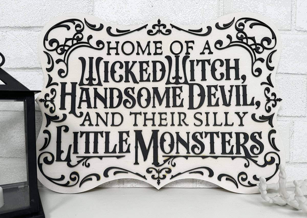 A decorative sign reads "Home of a wicked witch, handsome devil, and their silly little monsters" with ornate black lettering on a white background.