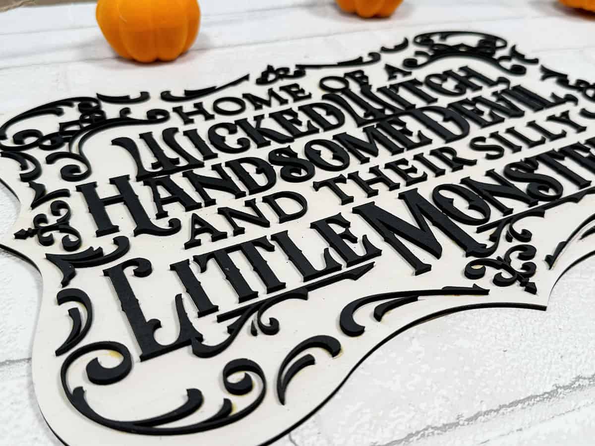 A decorative sign reads "Home of a Wicked Witch, Handsome Devil, and Their Silly Little Monster," with small pumpkins in the background on a white surface.