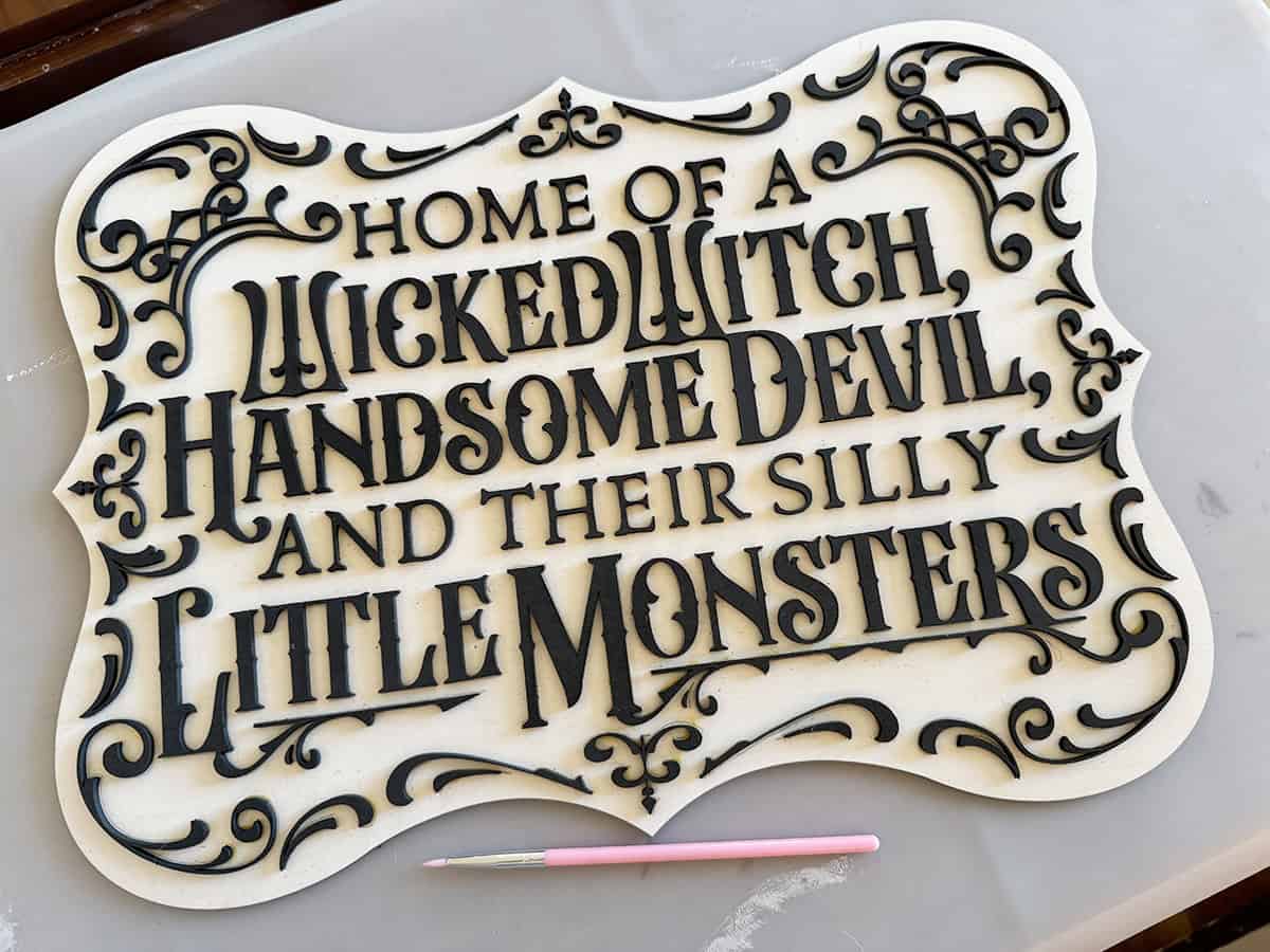 A decorative white sign with black text reads "Home of a Wicked Witch, Handsome Devil, and their Silly Little Monsters," placed on a table with a small pink tool next to it.