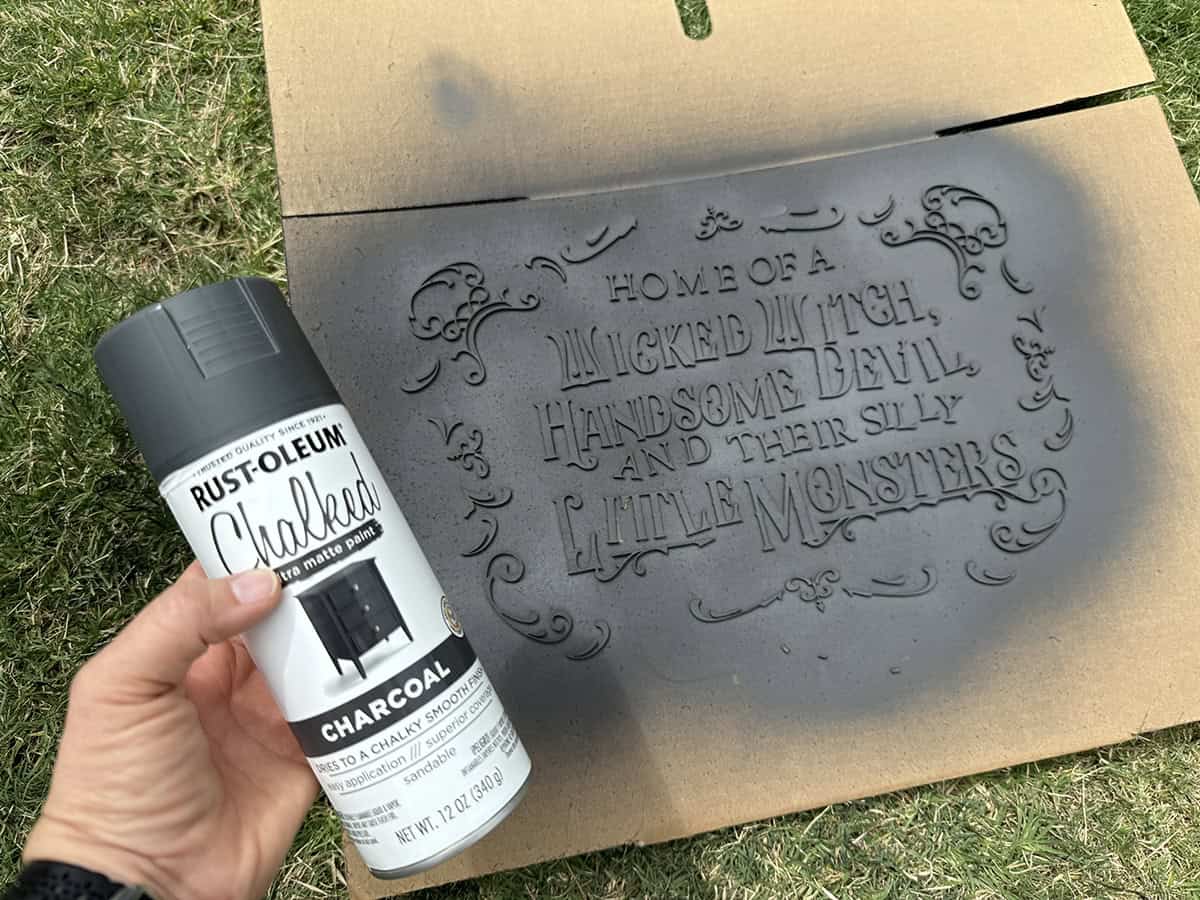A hand holds a can of Rust-Oleum Chalked spray paint over a stencil reading "Home of a wicked witch, handsome devil, and their silly little monsters" on cardboard with a grass background.