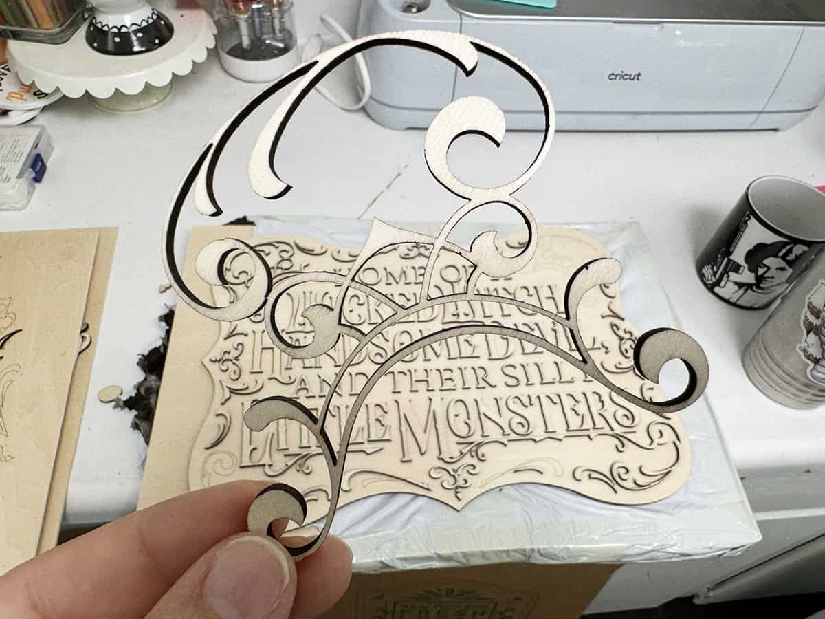 A hand holds a delicate wooden cutout with a swirl design. In the background, a laser-cut sign with text and a craft machine are visible.