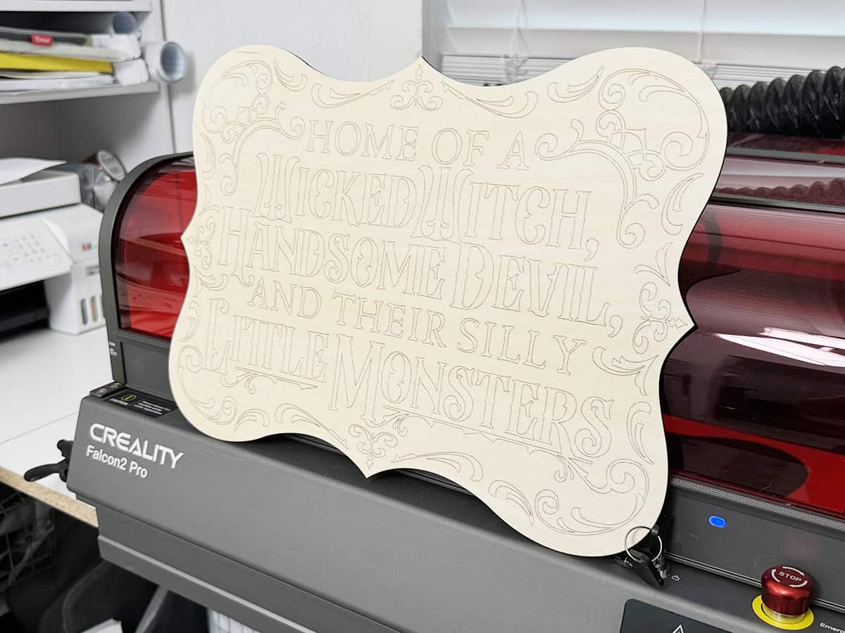 A decorative wooden sign with ornate edges and engraved text that reads, "Home of a wicked witch, handsome devil, and their silly little monsters," placed on a Creality Falcon2 Pro machine.
