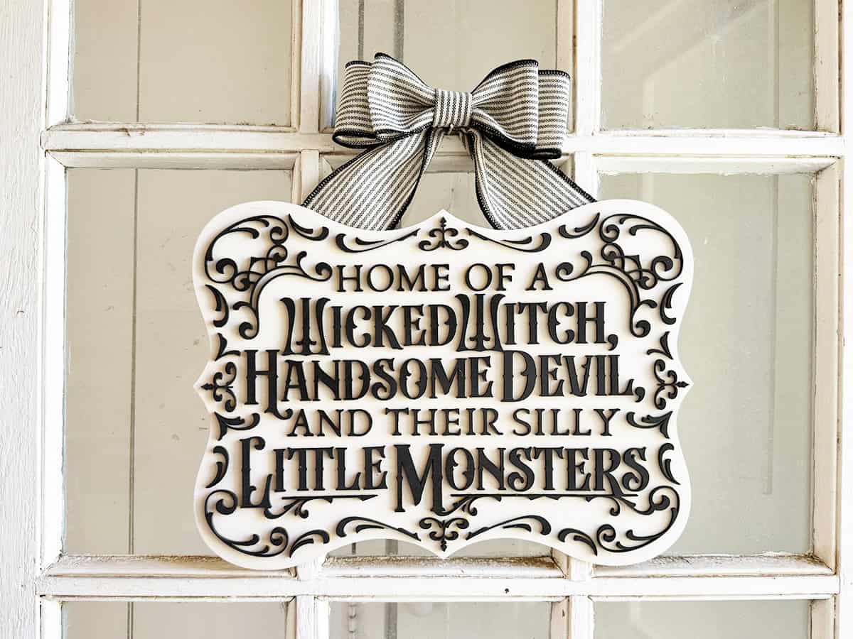 A decorative sign on a door reads, "Home of a Wicked Witch, Handsome Devil, and Their Silly Little Monsters," with a bow at the top.