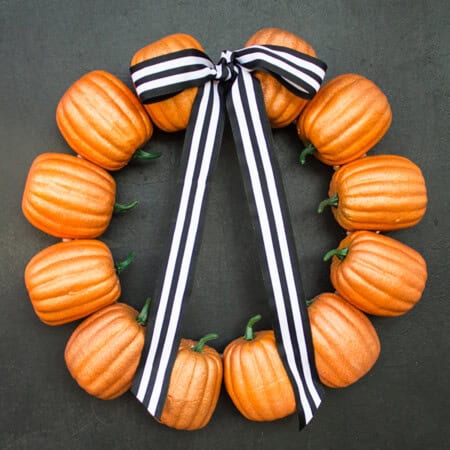 A Halloween Pumpkin Wreath made of small orange pumpkins from Dollar Tree, each with a green stem, arranged in a circle on a dark background. A black and white striped ribbon is tied in a bow at the top.