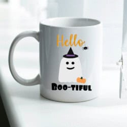 White mug with a ghost illustration, wearing a witch hat, and holding a small pumpkin. Text reads "Hello Boo-tiful" with a small spider beside it.