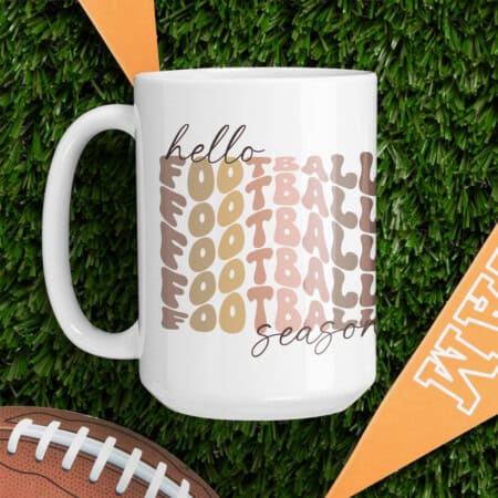 A white mug on green artificial turf with "hello football season" text in various shades of brown. A football and orange flags are also in the scene, perfect for anyone looking to download a football free SVG file to enhance their game day decor.