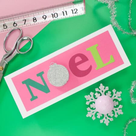A craft desk with a "Noel" card featuring colorful letters, a ruler, scissors, silver tinsel, and snowflake decorations on a green surface.