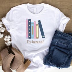 White t-shirt with pastel-colored illustrated books and the text "I'm booked!" laid on a burlap surface with jeans, brown boots, and cotton stems.