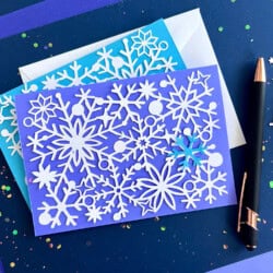 A blue greeting card with intricate white snowflake cutouts, accompanied by an envelope and a black pen on a dark blue background scattered with colorful confetti.