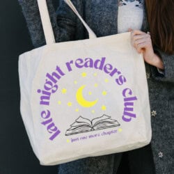 A person holds a tote bag with the text "Late Night Readers Club" and an illustration of a book and moon with stars.