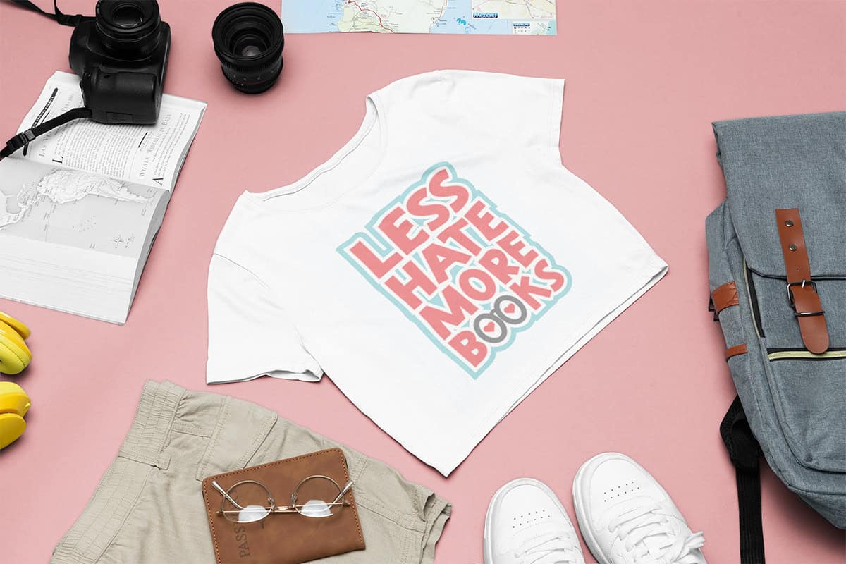 A white t-shirt with "Less Hate More Books" text lies on a pink surface, surrounded by a camera, map, shorts, passport, headphones, shoes, and a backpack.