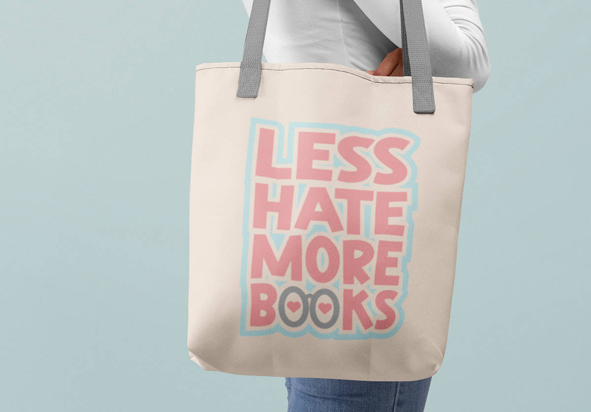 A person holding a beige tote bag with gray straps displaying the text "Less Hate More Books" in pink and blue.