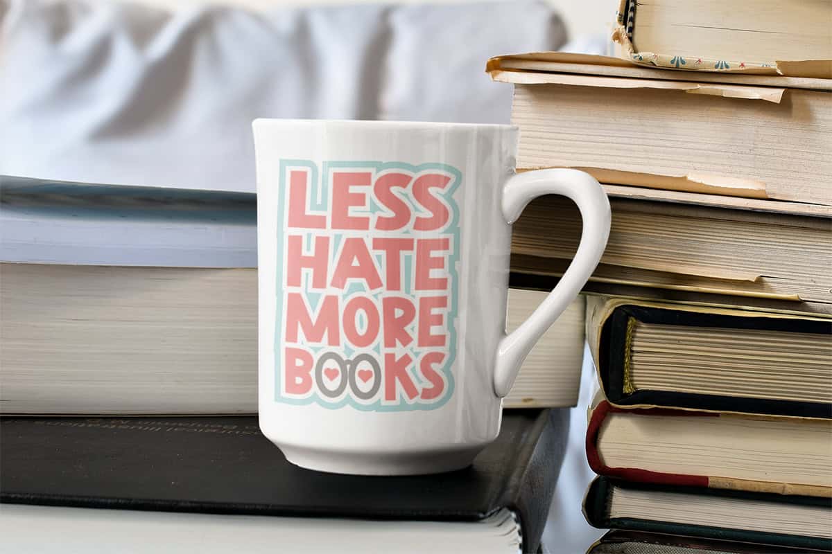 A white mug with "Less Hate More Books" printed in pastel colors is placed beside a stack of books.