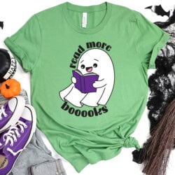Green t-shirt with a cartoon ghost reading a book and the text "read more boooooks." Nearby are purple sneakers, a small pumpkin, and Halloween decorations.