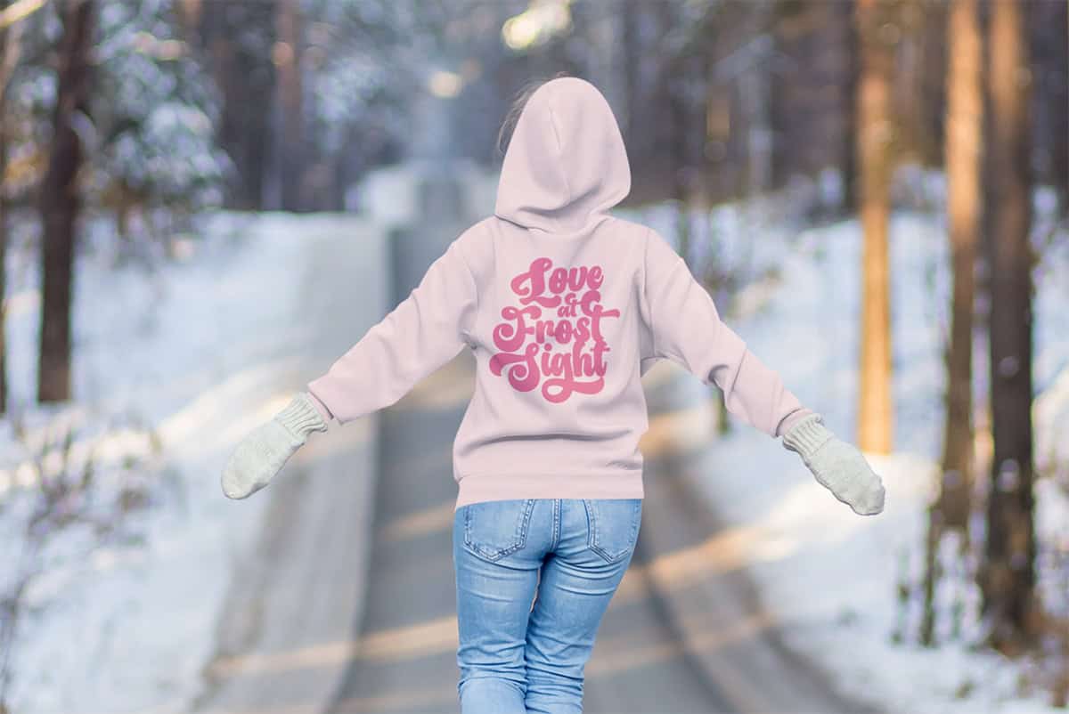Person in a pink hoodie with "Love at First Sight" text, walking on a snowy road between trees.