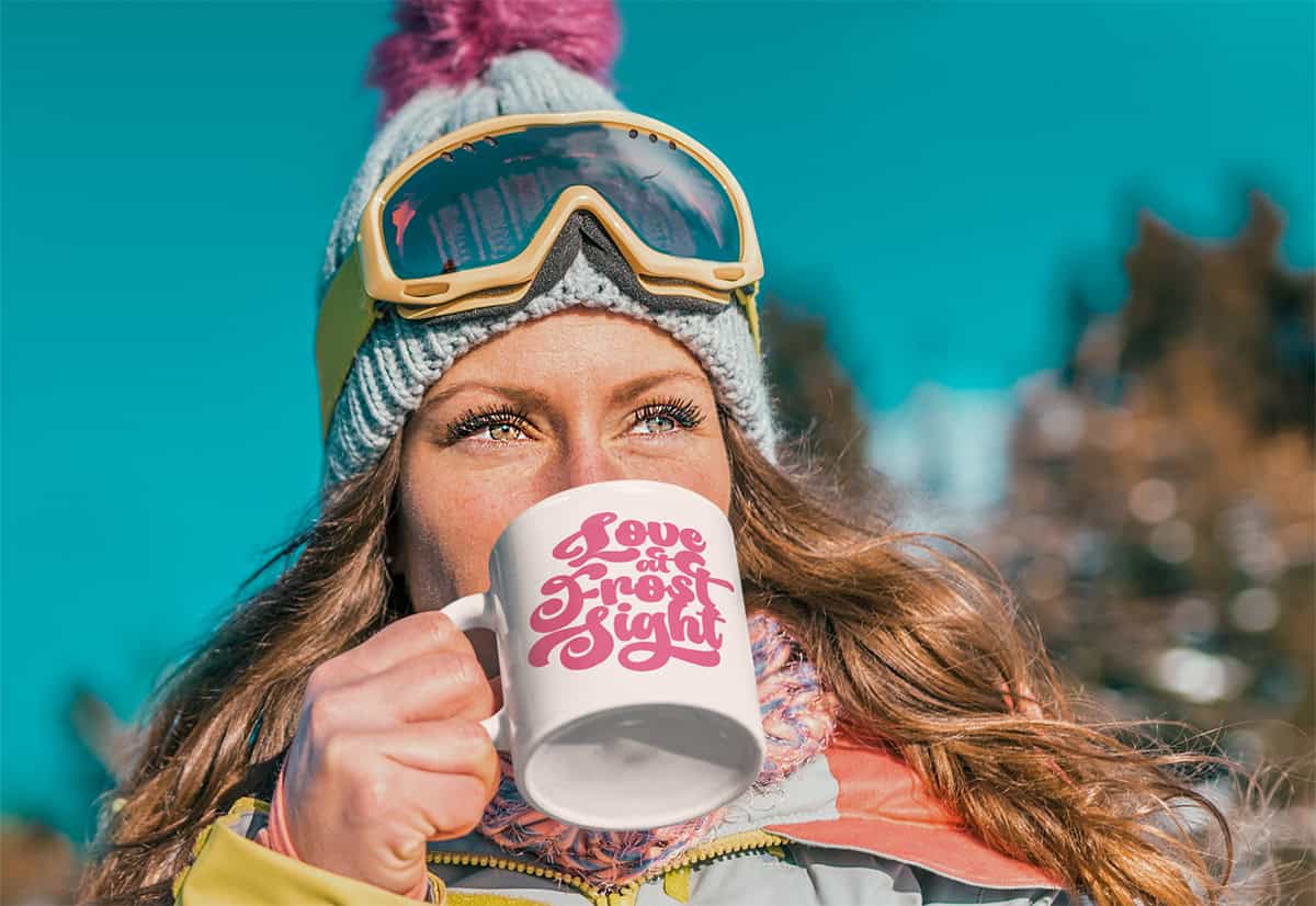 A person in winter clothing and goggles drinks from a mug with "Love at Frost Sight" printed on it, against a snowy background.
