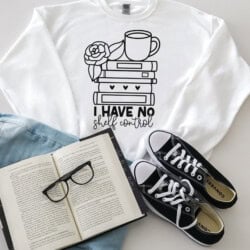 White sweatshirt with "I Have No Shelf Control" design featuring books, a rose, and a mug. Surrounded by open book, black glasses, denim jeans, and black sneakers.