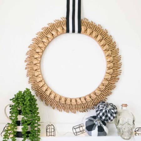 A decorative wreath with a golden acorn design hangs on a striped ribbon. Below it are mini pumpkins wrapped in black and white cloth, a skull ornament, a potted plant, and wireframe lights.