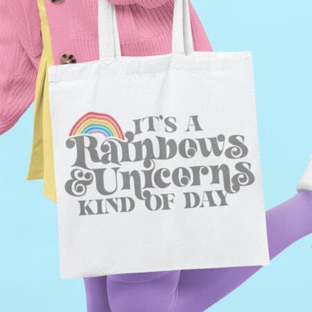 A person holding a white tote bag with the text "It's a Rainbows & Unicorns Kind of Day" and a rainbow graphic. The person, dressed in colorful clothing, epitomizes the joy of finding that perfect unicorn free SVG design.