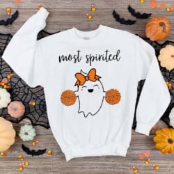 White sweatshirt with a cute ghost illustration holding pom-poms, wearing an orange bow. The text "most spirited" is above. Surrounded by pumpkins, candy corn, and bat decorations on a wooden surface.