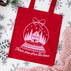 Red tote bag with a white snow globe design featuring a house and trees, and the text "There's snow place like home" surrounded by festive decorations.