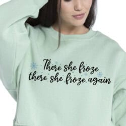 Woman wearing a light green sweatshirt with the text "There she froze, there she froze again" and small snowflake designs.
