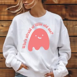 Person wearing a white sweatshirt with a smiley ghost graphic and the text "Who's afraid of little old me?" in red.